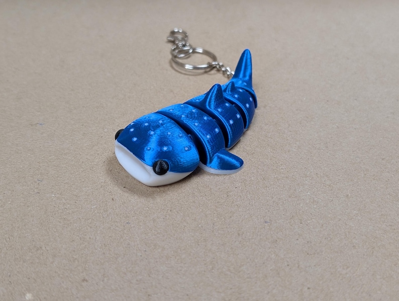 May include: A blue and white articulated whale shark keyring featuring a segmented body, a flat white underside, and small white spots across the top. The shark is attached to a silver-coloured metal split ring and a short chain for keys.