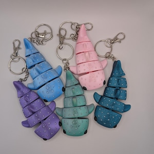 May include: Five articulated whale shark keychains in purple, light blue, teal, pink, and dark blue. Each keychain features a segmented body with a spotted pattern, small fins, and a metal lobster claw clasp attached to a circular split ring.
