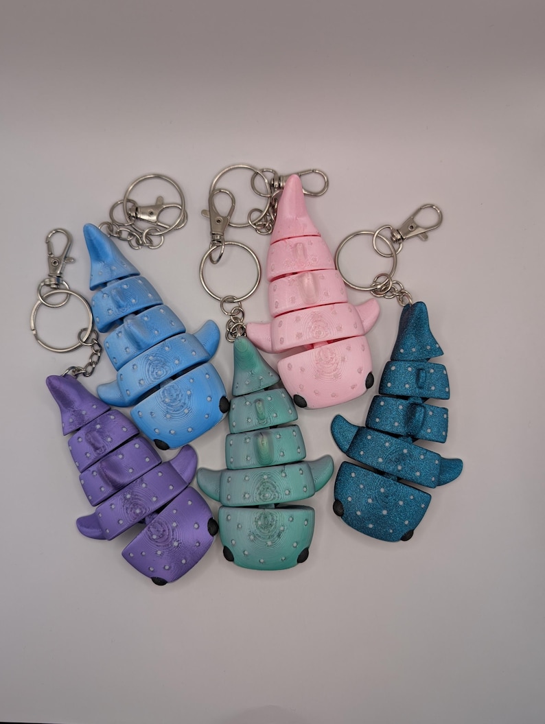 May include: Five articulated whale shark keychains in purple, light blue, teal, pink, and dark blue. Each keychain features a segmented body with a spotted pattern, small fins, and a metal lobster claw clasp attached to a circular split ring.