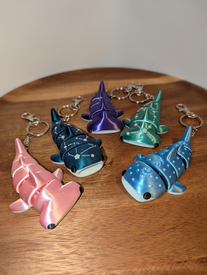 May include: Five articulated shark keychains in pink, dark blue with white star patterns, purple, teal with white star patterns, and light blue with white polka dots. Each shark features a metal keyring and lobster clasp attachment.
