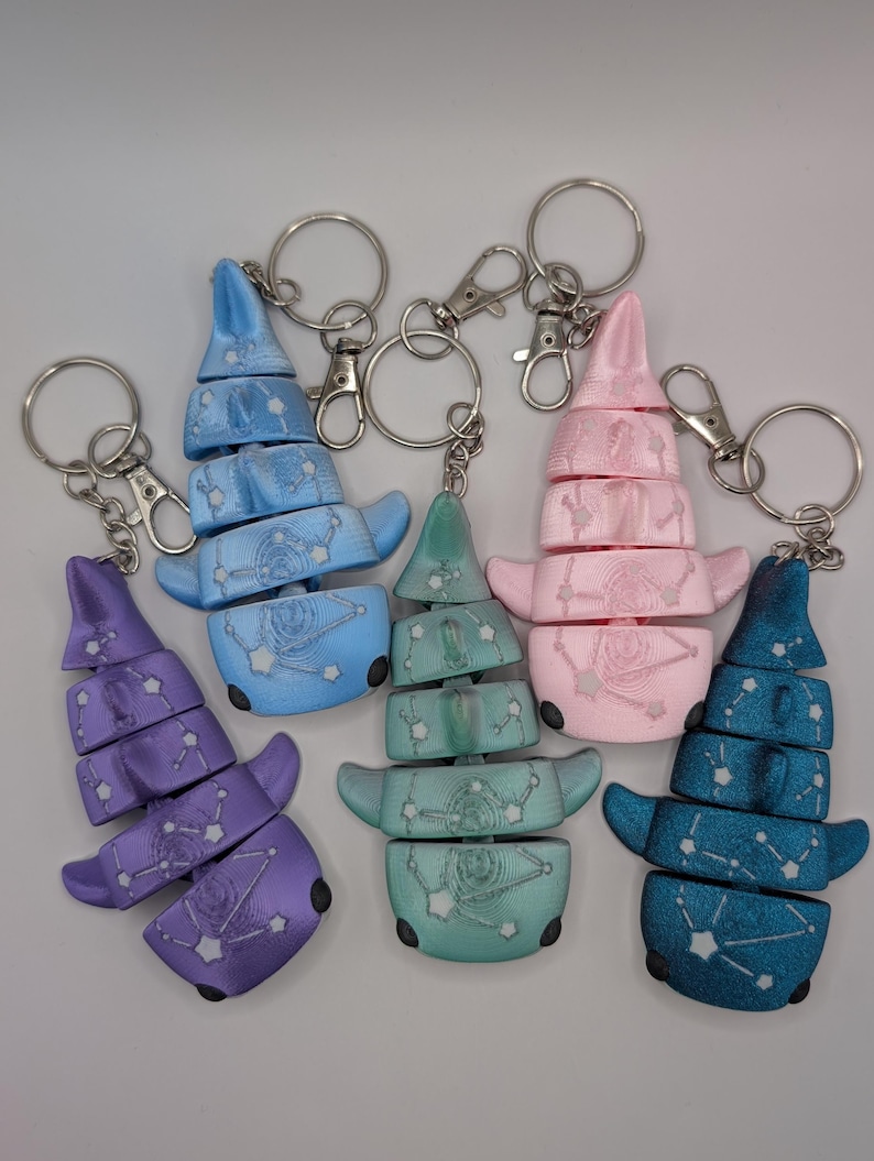 May include: Five articulated shark keychains in purple, light blue, teal, pink, and dark blue. Each shark features a 3D-printed texture with a white constellation pattern across the body, a segmented design, and a silver metal keyring attachment.