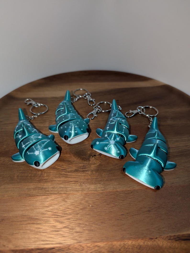 May include: Four articulated shark keychains in a metallic teal finish. Two are whale sharks and two are hammerhead sharks, each featuring white constellation patterns on their backs, black eyes, and silver-toned metal lobster claw clasps.