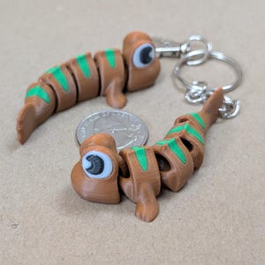 May include: Two brown and green articulated slug-like figures, one attached to a silver metal keychain. Each figure features large white eyes with black pupils. A United States quarter coin is placed between the two figures for scale.