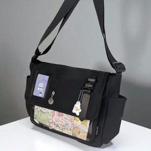 May include: Black messenger bag with a flap closure and adjustable shoulder strap. The bag features a clear pocket displaying a colourful floral design and a small charm. The bag has a side pocket and a small tag that says "POPPY HT".