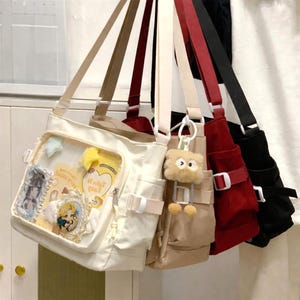 Puede incluir: Una colección de bolsos de hombro en varios colores, incluyendo blanco, beige, rojo y negro. El bolso blanco tiene un bolsillo transparente lleno de objetos decorativos. Cada bolso tiene una correa ajustable y un bolsillo. Un pequeño llavero esponjoso cuelga de un bolso.