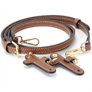 Pode incluir: Alça de bolsa de couro marrom com ferragens douradas. A alça é feita de couro marrom texturizado com costura branca e inclui dois fechos giratórios dourados e duas peças de conexão de couro.