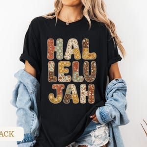 May include: A black short-sleeved t-shirt featuring the word HALLELUJAH printed in large, blocky letters. Each letter is filled with a multi-colored floral pattern in shades of orange, yellow, brown, and cream, creating a distinct contrast against the black fabric.