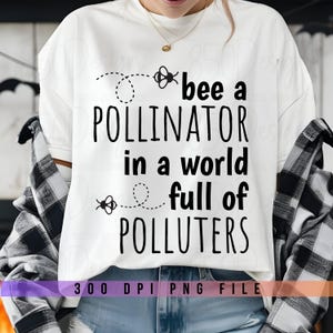 Op de afbeelding: Wit T-shirt met zwarte tekst die "bee a pollinator in a world full of polluters" luidt. De tekst wordt omringd door kleine bij-illustraties. Onderaan het shirt staat "300 DPI PNG FILE".