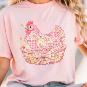 May include: A light pink t-shirt with a watercolour illustration of a pink hen in a woven basket. The hen has a floral and polka dot pattern. The basket has a bow on the front.