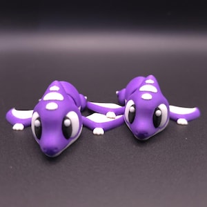 May include: Two purple dragon figurines with white accents. The dragons have large black eyes and are lying down. The figurines are on a dark gray surface.