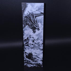 Peut inclure: Une illustration en noir et blanc d'un dragon volant au-dessus d'une chaîne de montagnes enneigées. La tête du dragon est au premier plan, ses ailes déployées derrière lui. Les montagnes sont en arrière-plan, avec des sommets enneigés.