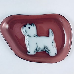 Può includere: Un piatto di vetro rosa con una statuetta di cane in ceramica bianca al centro. Il cane è rivolto verso il lato sinistro del piatto e ha occhi neri e un naso nero.