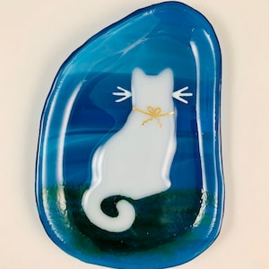 Op de afbeelding: Een wit glazen kattenbeeldje met een gouden strik zit in een blauwe glazen schaal met een groene swirl-ontwerp.