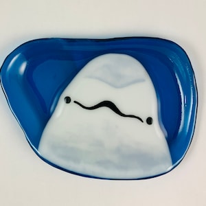 Op de afbeelding: Een blauwe glazen schaal met een wit, gestileerd dolfijnontwerp. De dolfijn heeft zwarte ogen en een mond.