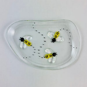Op de afbeelding: Een heldere glazen schaal met een afgeronde vorm en drie erop geschilderde gele en zwarte bijen. De bijen vliegen in verschillende richtingen en zijn omringd door zwarte stippen.