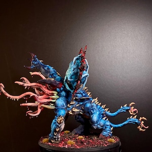 May include: A blue, multi-limbed creature figurine with a central mass of pink, tentacle-like appendages extending from its torso. The figure features sharp, horn-like protrusions, a large dorsal fin, and two long, thin tails ending in claws.