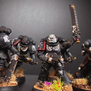 Op de afbeelding: Miniatuurfiguren van Space Marines in zwarte bepantsering, met wapens. De figuren staan op een bruine basis met miniatuurplanten. Eén figuur houdt een groot, gekarteld mes vast. De bepantsering heeft witte en rode accenten.