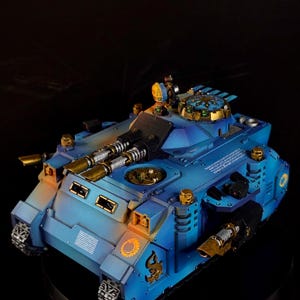Puede incluir: Maqueta detallada de un tanque azul y dorado. El tanque presenta cañones dobles, detalles intrincados y detalles dorados. El modelo está sobre un fondo oscuro, resaltando su diseño y artesanía.