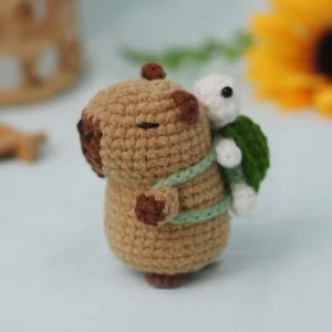 May include: A brown crocheted capybara with a white crocheted turtle on its back. The turtle is wearing a green shell and has a white face.