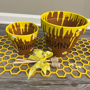 May include: Two brown terracotta flower pots with yellow paint dripping from the rim, featuring the word "HUNNY" in black lettering and small bee embellishments. Included are two wooden honey dippers and a yellow ribbon with a bee pattern.