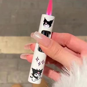 May include: A white lighter with black cartoon character graphics and pink flame. The lighter features a button and a decorative design of diamonds. The lighter is held in a hand.