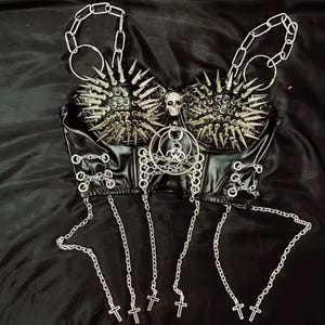 May include: Black faux leather bustier top with silver metal spikes, chains, and crosses. The top features a skull and a biohazard symbol. The chains hang from the bottom and the top of the bustier.