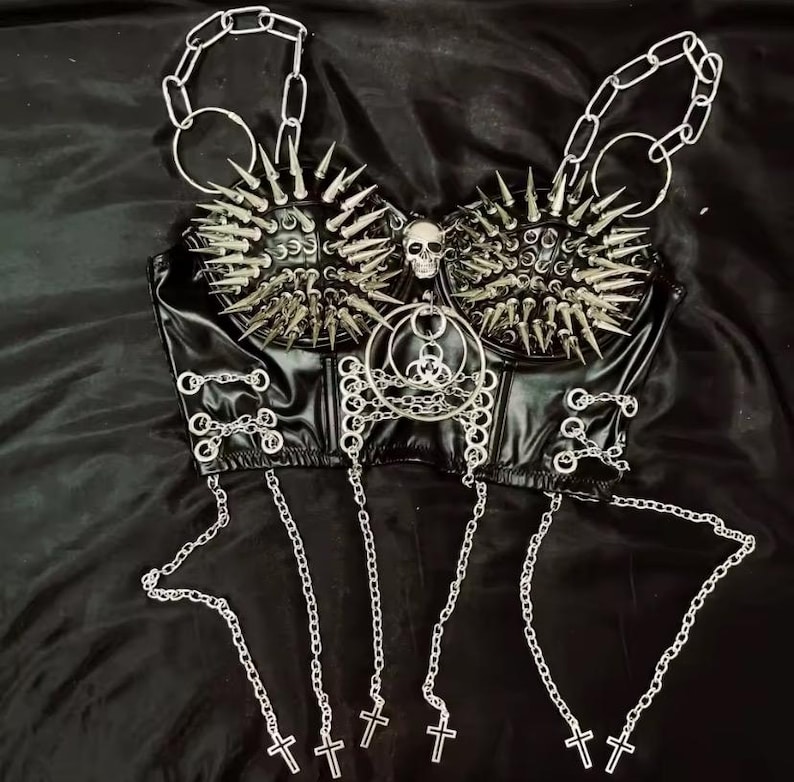 May include: Black faux leather bustier top with silver metal spikes, chains, and crosses. The top features a skull and a biohazard symbol. The chains hang from the bottom and the top of the bustier.