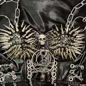 May include: Black faux leather corset with silver metal spikes, chains, and a skull pendant. The corset features a structured design with multiple rows of spikes and chain detailing, creating a bold, edgy aesthetic.