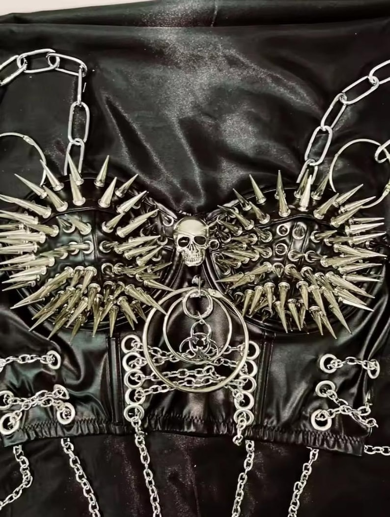 May include: Black faux leather corset with silver metal spikes, chains, and a skull pendant. The corset features a structured design with multiple rows of spikes and chain detailing, creating a bold, edgy aesthetic.
