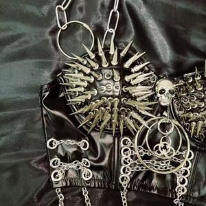 May include: Black faux leather corset top with silver metal spikes, chains, and a skull accent. The corset features a studded design and multiple chain details, creating a bold, edgy aesthetic. The top is designed with a structured silhouette.