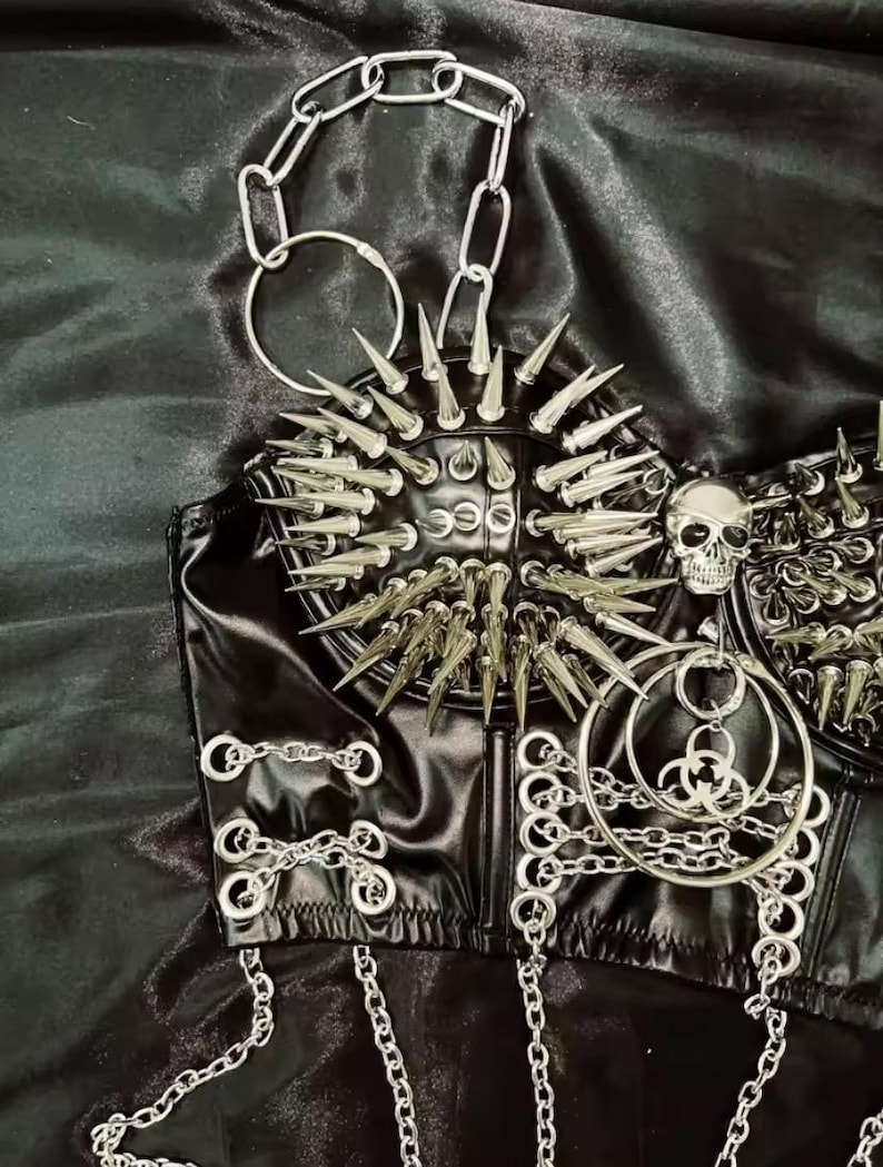 May include: Black faux leather corset top with silver metal spikes, chains, and a skull accent. The corset features a studded design and multiple chain details, creating a bold, edgy aesthetic. The top is designed with a structured silhouette.