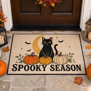 Peut inclure: Un paillasson rectangulaire illustré d'un chat noir assis devant un croissant de lune jaune, entouré de trois citrouilles orange et crème, de petites chauves-souris et d'étoiles, avec le texte SPOOKY SEASON en lettres noires grasses.