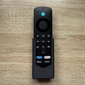 Op de afbeelding: Zwarte afstandsbediening met een aan/uit-knop, een cirkelvormig navigatiepad en diverse functieknoppen. De afstandsbediening heeft knoppen voor Prime Video, Netflix, Disney+ en Peacock. Geplaatst op een houten oppervlak.