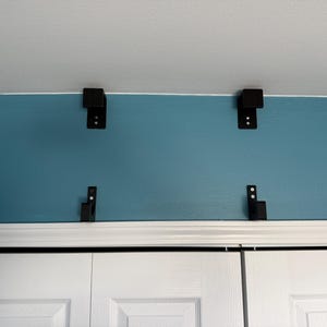 May include: Four black metal brackets are mounted on a teal wall, above a white cabinet. The brackets are evenly spaced and appear to be designed for supporting a shelf or other structure. The brackets are rectangular with two visible screw holes.