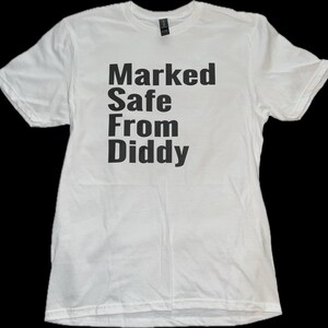 May include: White t-shirt with the phrase "Marked Safe From Diddy" printed in bold black letters. The shirt is made of a soft material and has short sleeves. The text is centered on the front of the shirt.