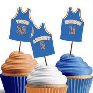 May include: Three cupcakes topped with blue basketball jersey-shaped paper toppers. The toppers feature the names TOWNS 32, ANUNOBY 8, and BRUNSON 11 in orange and white text. The cupcakes have orange, white, and blue frosting in blue and orange liners.