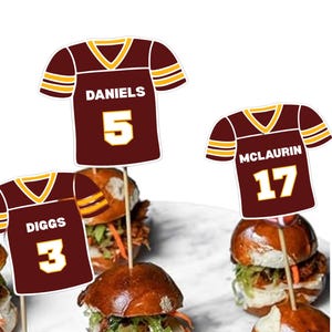 May include: Three burgundy football jersey-shaped cupcake toppers with yellow and white trim. Each features a player name and number: DANIELS 5, DIGGS 3, and MCLAURIN 17. The toppers are attached to thin wooden sticks for insertion into food items.