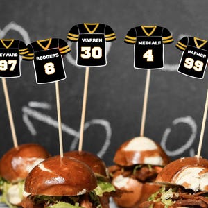 Könnte beinhalten: Miniatur-Football-Trikot-Food-Picks auf Mini-Burgern. Die schwarzen und goldenen Trikot-Picks haben Spielernamen und -nummern. Die Mini-Burger sind mit Salat und Fleisch gefüllt, mit Brezelbrötchen.