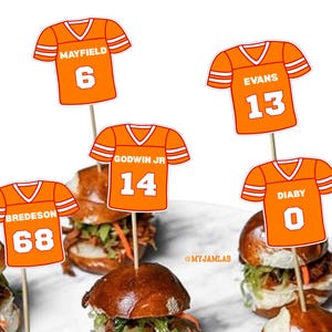 May include: Orange football jersey-shaped food picks with white stripes and numbers, atop mini sandwiches. The picks display names and numbers: Mayfield 6, Evans 13, Godwin Jr 14, Diaby 0, and Bredeson 68. The sandwiches have a dark bun and filling.
