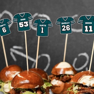May include: Small sandwiches with wooden skewers displaying football jersey designs. The jerseys are teal and white, with names and numbers such as "Smith 6" and "Hurts 1." The sandwiches have pretzel buns, lettuce, and meat filling.