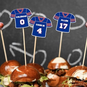 May include: Three blue football jersey cupcake toppers on wooden picks. The jerseys feature red and white trim with names and numbers: COLEMAN 0, COOK III 4, and ALLEN 17. These paper cutouts are inserted into small pretzel bun sliders.