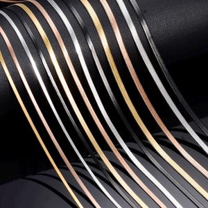 Könnte beinhalten: Eine Sammlung flacher Fischgrätenketten, die in parallelen Linien angeordnet sind. Die Ketten haben eine metallische Oberfläche und sind in verschiedenen Farben wie Gold, Silber, Roségold und Schwarz erhältlich und zeigen eine gewebte Struktur.