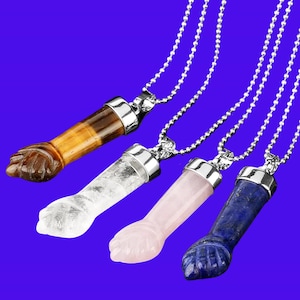 May include: Four gemstone fist pendants on silver-tone chains. The gemstones are brown, clear, pink, and blue. Each pendant has a silver-tone metal cap. The background is a solid blue.