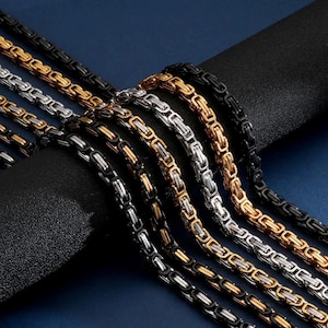May include: Close-up of several Byzantine chain necklaces in various colors, including gold, silver, and black. The chains are arranged in a layered pattern, showcasing their intricate design and metallic sheen. The background is a dark blue.