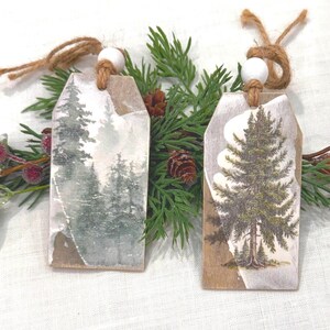 May include: Two rustic Christmas tree-shaped gift tags with forest scenes. One tag features a misty forest, the other a detailed evergreen tree. Each tag has a jute string and a white bead for hanging. The tags are surrounded by greenery.