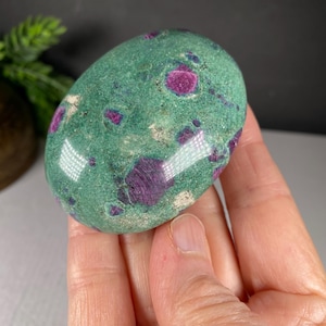 May include: A polished oval-shaped stone featuring a vibrant green base color with embedded deep magenta and purple crystalline inclusions throughout the surface. The stone has a smooth, glossy texture and displays natural mineral variations.