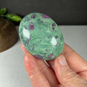 May include: An oval-shaped polished stone featuring a vibrant green base color with scattered, deep magenta-colored inclusions throughout the surface. The stone has a smooth, glossy texture and exhibits natural variations in its mineral composition.