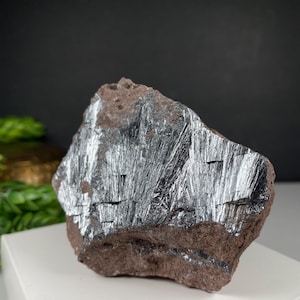 May include: A raw, natural mineral specimen featuring a dark, metallic silver-gray surface with a striated texture. The mineral is set in a reddish-brown matrix, creating a striking contrast. The overall shape is irregular and rough.