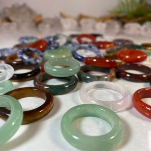 May include: A collection of colorful gemstone rings. The rings are made of various stones, including green, red, brown, and white. The rings are all different sizes and shapes.
