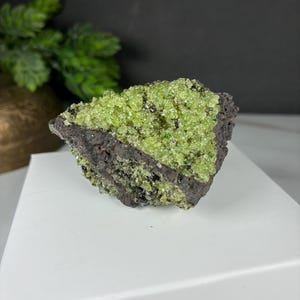 May include: A raw mineral specimen featuring a cluster of vibrant lime green crystals embedded within a dark, porous volcanic rock matrix. The crystalline surface displays a granular texture with varying shades of bright green and black inclusions.